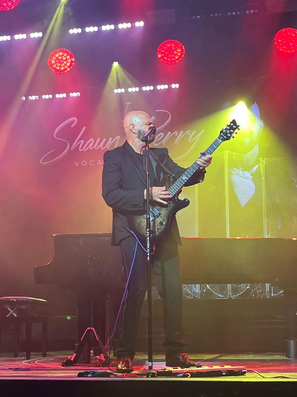 Shaun Perry performing with guitar on a colourful theatre stage